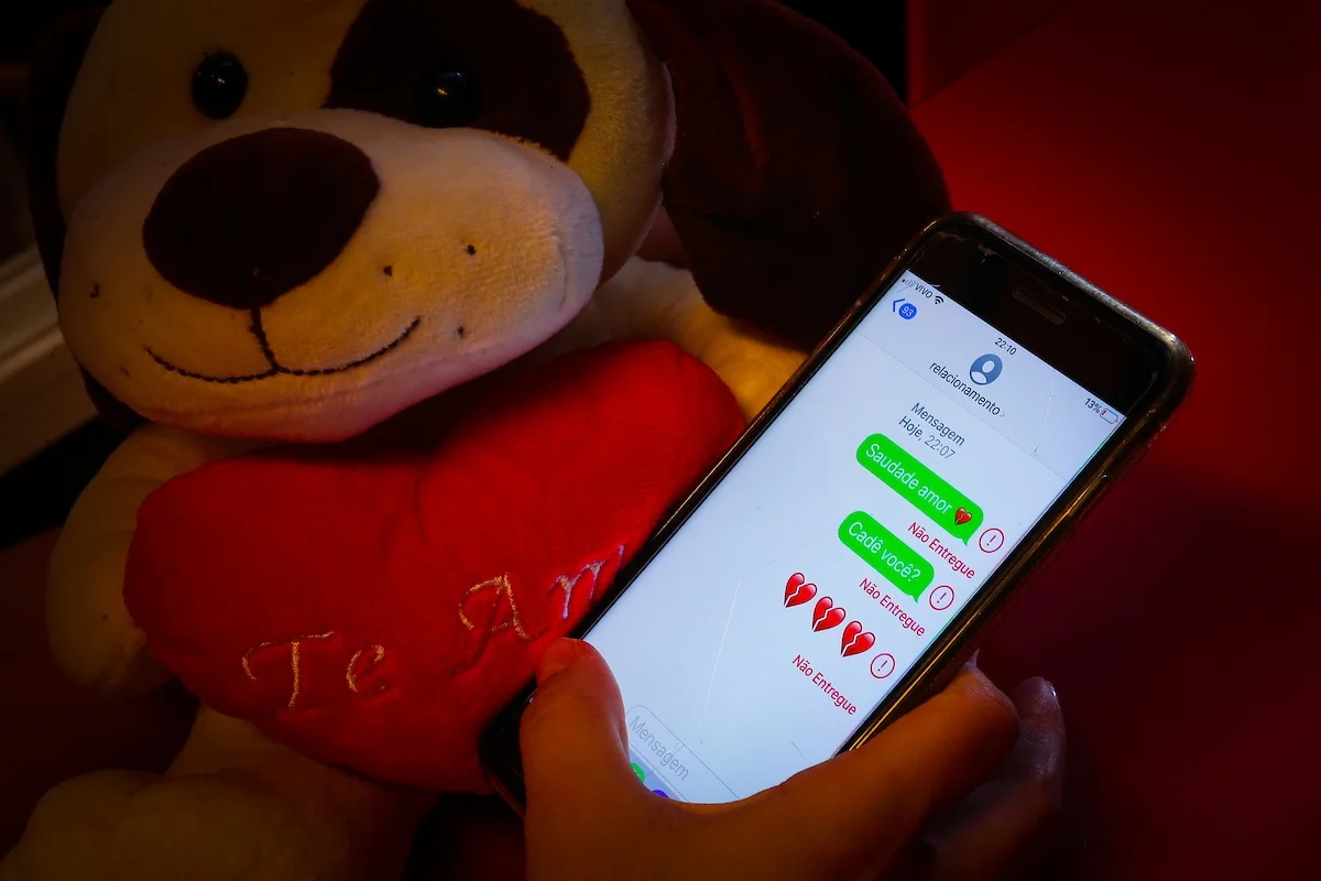 Imagem colorida de um urso, um coração vermelho, escrito 'Te Amo', uma mão segurando um celular com mensagens referentes a relacionamento