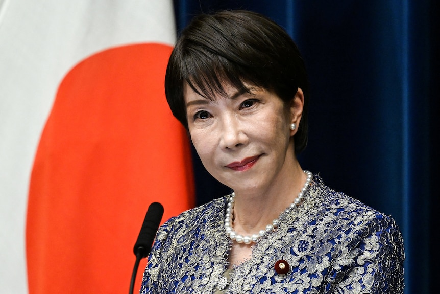 Takaichi Sanae with a serious expression stands in front of a Japanese flag.